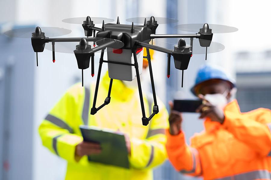 A drone flying in the air with two people dressed in orange and yellow work jumpsuits are in background